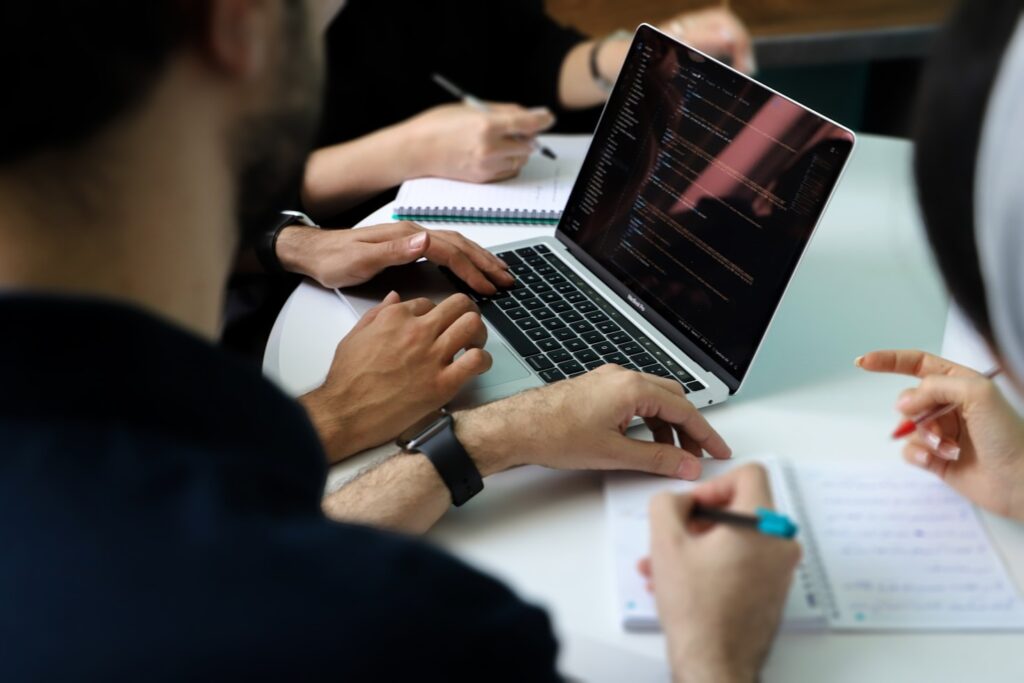 Hands working at a laptop surrounded by others at a meeting table.